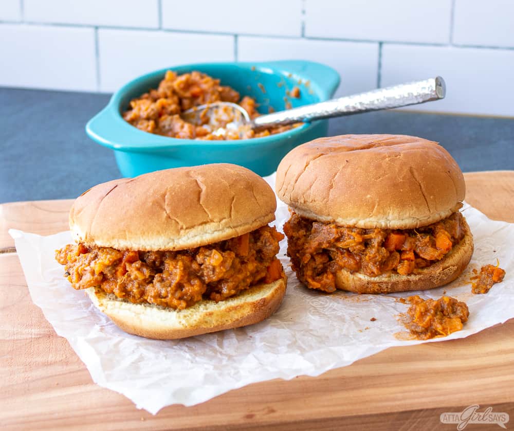 two sloppy Joes sandwiches on a wooden board with additional filling in a pan in the background