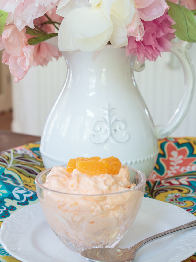 Orange Fluff dessert in a clear glass bowl with a pitcher of flowers