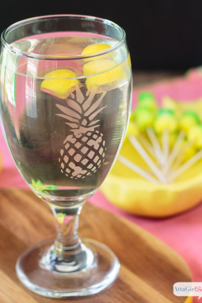 A pineapple drinking glass filled with water and pineapple juice ice cubes. The water glass features an etched pineapple on it and sits atop a wooden cutting board. There's a yellow bowl filled with pineapple cocktail picks in the background.