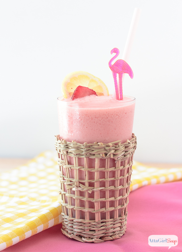 A tall glass of frozen strawberry lemonade garnished with lemon and strawberry slices. Served in a beachy style glass with a pink straw and a flamingo cocktail pick.