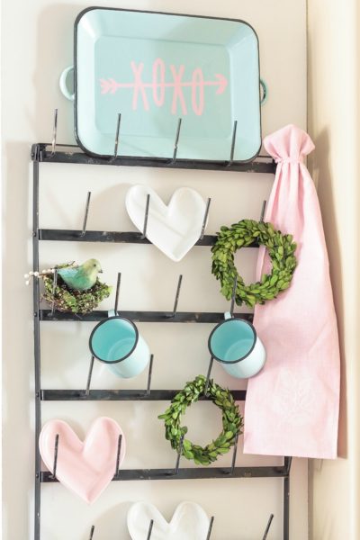 Dress up your coffee mug wall rack with simple Valentine's Day decor, including a "Gossip Girl" inspired quote you can make with your Cricut. Enamel coffee mugs, miniature boxwood wreaths and heart-shaped plates! Swoon. Click for sources to recreate this look! #mugrack #valentinesday #gossipgirl #xoxo #farmhouse #aquaandpink #mugrackmonday #valentinesdecor #cricutmade #cricutmaker #cricutexploreair #quotes