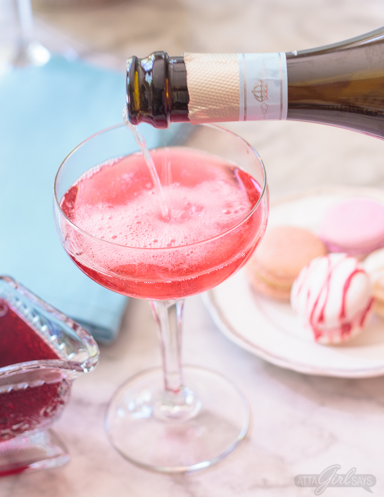 pouring prosecco into a hibiscus cocktail in a coupe glass