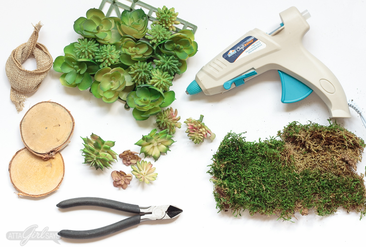 A collection of supplies required to make a DIY succulent napkin ring. The photo shows a glue gun, sheet moss, faux succulent plants, wire cutters, birch wooden round coasters and burlap jute ribbon.
