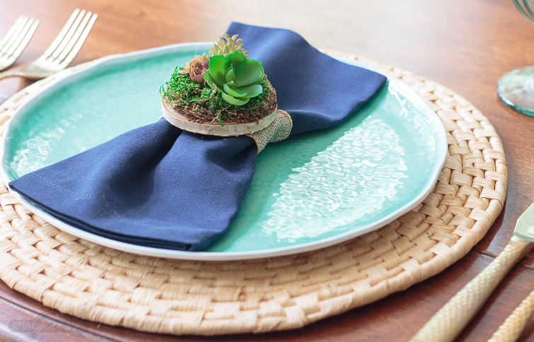 Dinner place setting featuring an aqua blue plate on a woven natural charger. A miniature succulent garden napkin ring holds a navy blue napkin