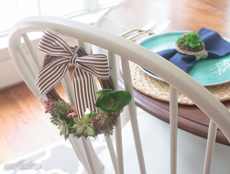 A white chair with a succulent grapevine wreath tied to the back of it. In the background, there's an aqua blue plate on top of a woven natural charger. A navy blue napkin, tied with a DIY succulent napkin ring miniature dish garden, rests on top of the plate.