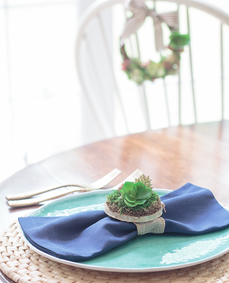 A dinner table place setting featuring an aqua blue plate on a woven seagrass charger. There's a navy blue napkin on the plate with a miniature succulent garden napkin ring. A grapevine succulent wreath hangs on a chair in the background.