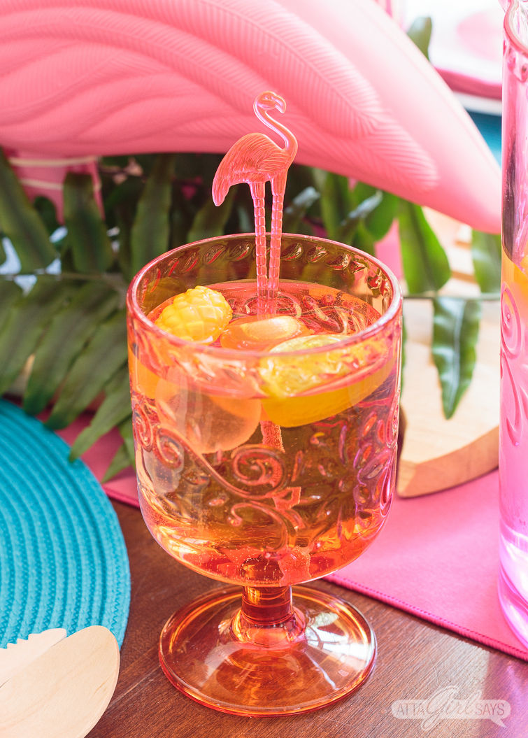 An orange plastic water goblet filled with colorful plastic fruit ice cubes and a flamingo swizzle stick.
