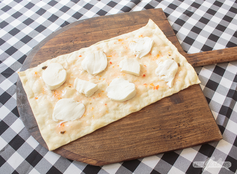 rectangular flatbread pizza crust, topped with pepper jelly and fresh mozzarella, on a wooden pizza peel on top of a gingham check tablecloth