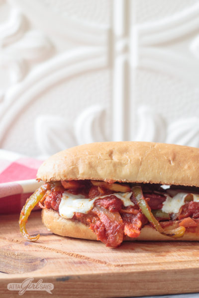 half of an Italian sausage and peppers sub sandwich covered in marinara sauce, sitting on a wooden cutting board with a red and white check napkin in the background left