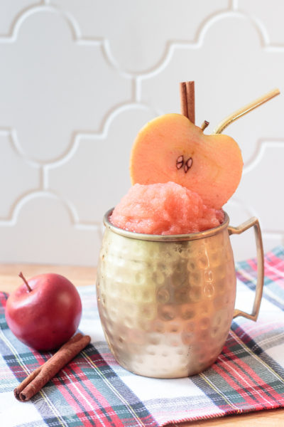 frozen apple cider cocktail slushie, garnished with an apple slice and cinnamon stick, in a gold Moscow mule mug. The cocktail is sitting on a green, white and black plaid napkin with a crabapple and cinnamon stick on the side