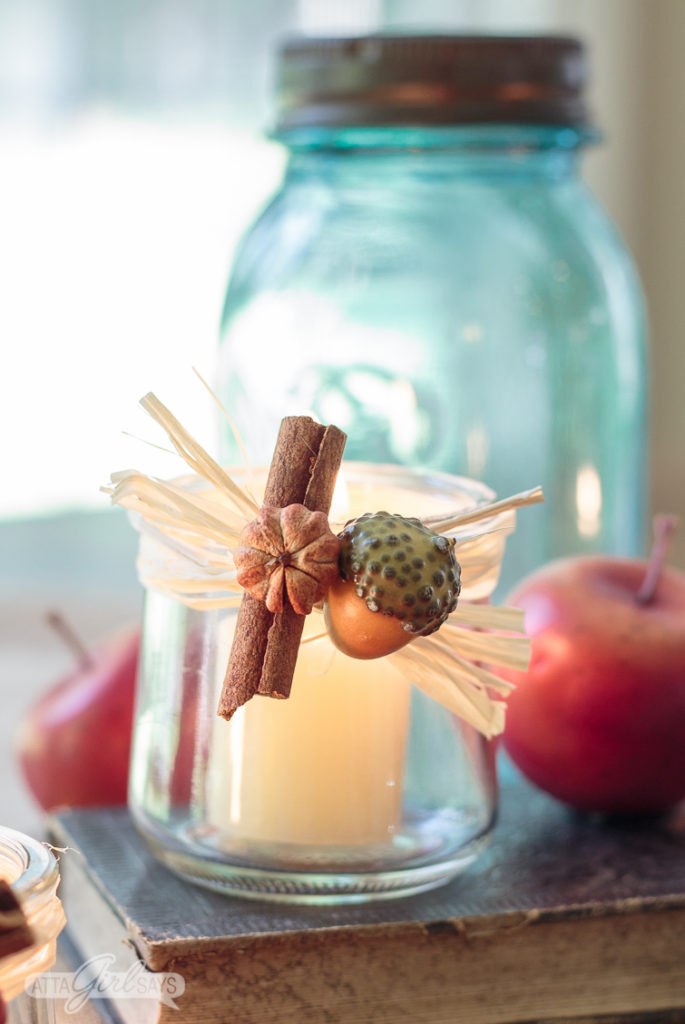 closeup of a glass yogurt jar candleholder decorated with miniature seed pods, acorn, raffia and a cinnamon stick. The votive is sitting on an old book with a blue glass mason jar and crabapples in the background