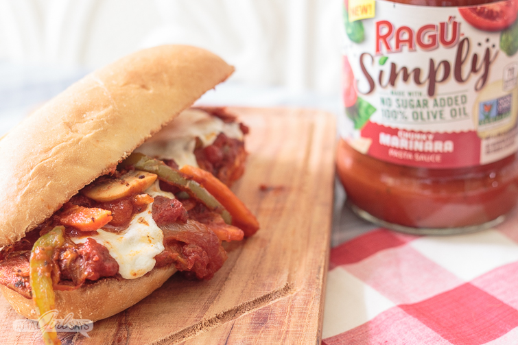 hot italian sausage and peppers sandwich on a wooden cutting board atop a red-and-white checked tablecloth with a jar of RAGU pasta sauce in the background.