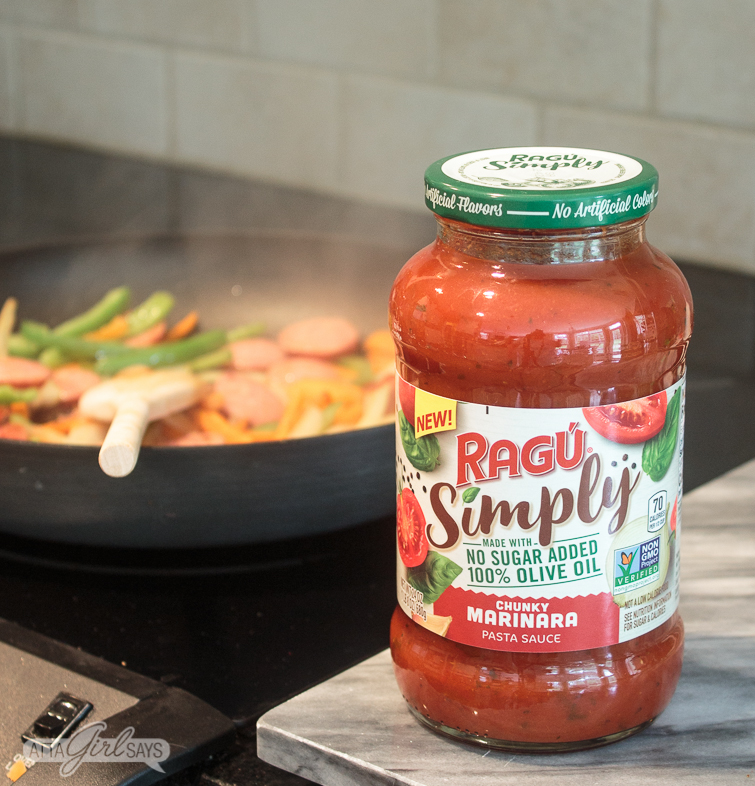 New RAGÚ&reg; Simply Chunky Marinara Pasta Sauce on a marble cutting board beside a stovetop with a pan of smoked sausage and vegetables in the background
