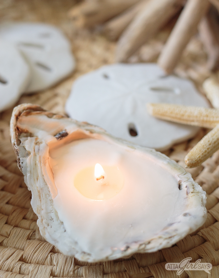 Add your favorite beach fragrance to these DIY oyster shell candles. How pretty would these be on the tables at a beach wedding or a romantic seaside dinner? #beachcrafts #seashellcrafts #oystershells #seashells #seashell #beachwedding #coastal #destinationwedding #diywedding #weddingdecor homemade oyster seashell candle burning on a seagrass mat with sand dollrs and starfish in the background