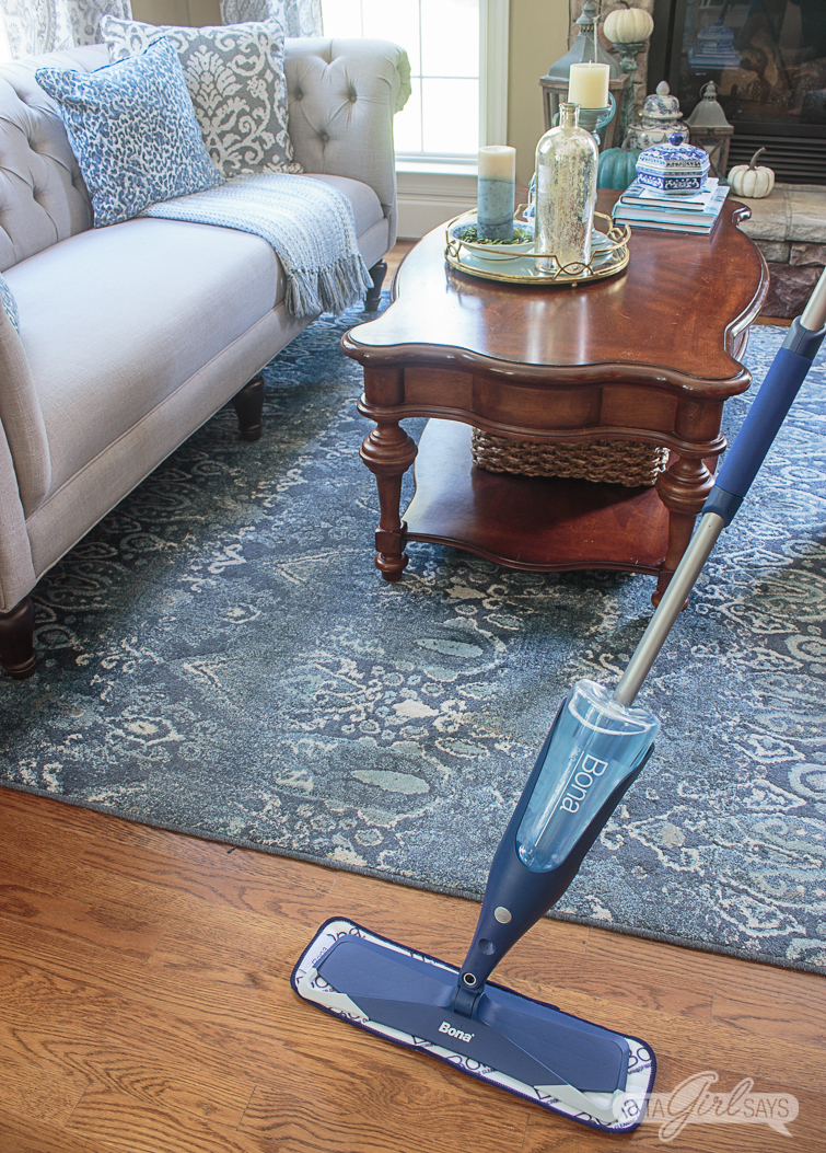using a Bona spray mop to clean hardwood floors in a living room with a blue rug and gray sofa in the background