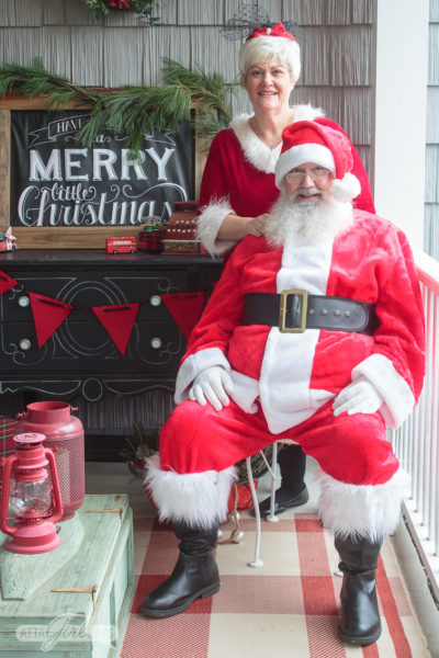 Santa and Mrs. Claus posing on a porch decorated in red, green and white vintage Christmas style