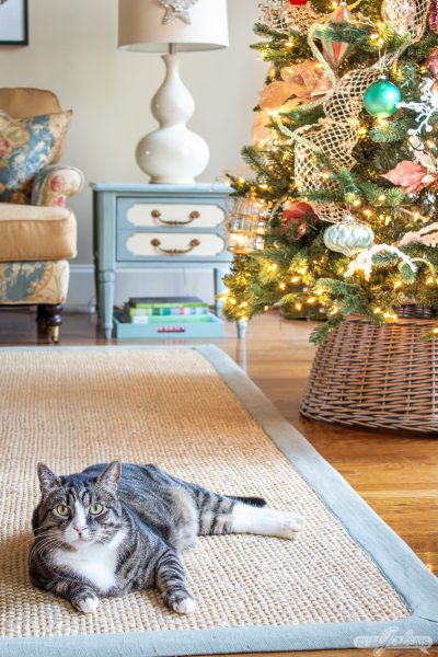 tabby cat laying on a seagrass rug in front of a Christmas tree
