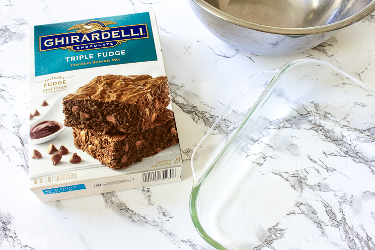 Ghiardelli triple fudge brownie mix, glass baking pan and a stainless steel bowl on a marble countertop