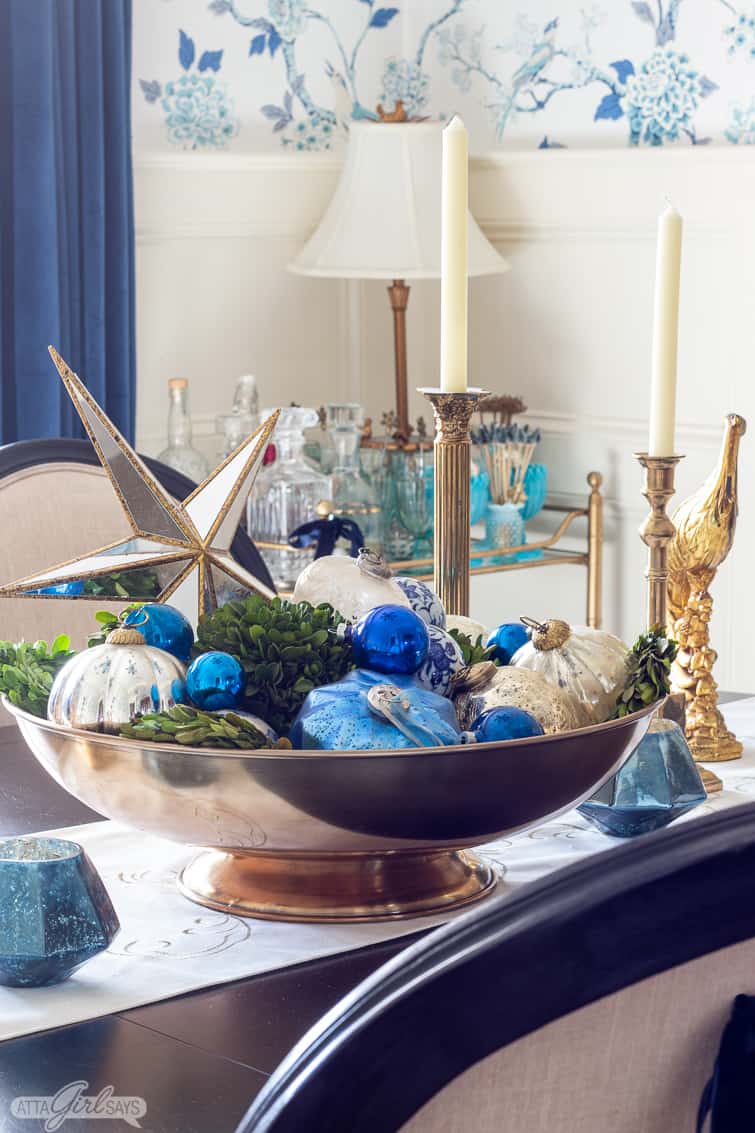 large brass centerpiece bowl filled with blue Christmas ornaments and boxwood balls on a dining room table