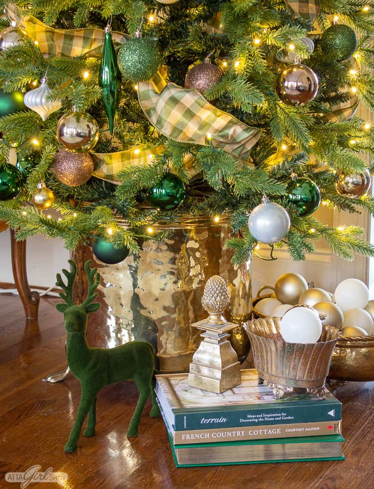 flocked deer, books and ornaments in brass containers underneath a Christmas tree in a brass planter