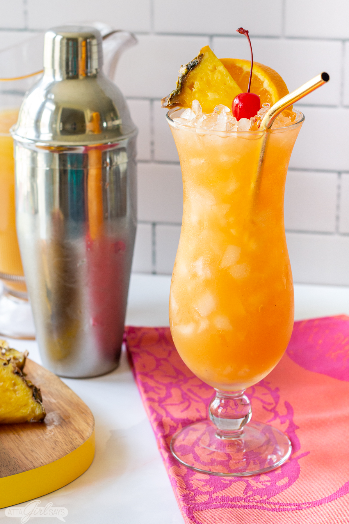 cocktail shaker and a Hurricane cocktail on a pink cloth with pineapple slices on a cutting board